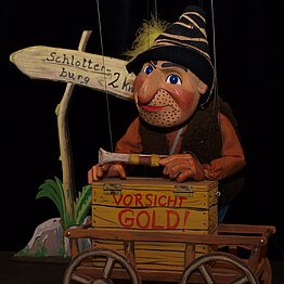 Eine Marionette aus Holz, sie stellt einen Mann mit runder Nase, roten Backen und Bartstoppeln dar. Er trägt eine Fellweste, ein orangenes Hemd und einen Räuberhut. In der Hand hat er eine Pistole. Vor ihm steht ein Wagen mit der Aufschrift "Vorsicht Gold!".