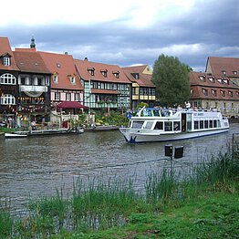 Ein Ausflugsschiff fährt auf einem Fluss, im Hintergrund Fachwerkhäuser.